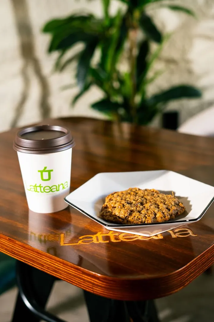 Café para llevar y galleta sobre mesa de madera con el logo de Latteana